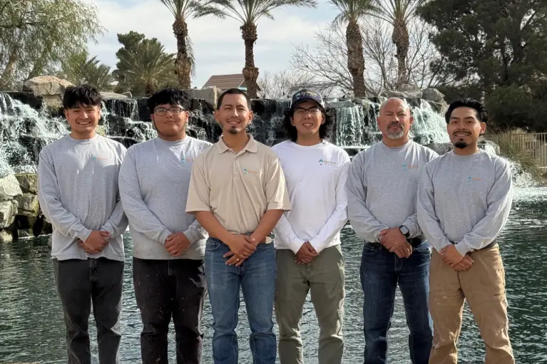 A group of six individuals stands by a serene waterfall and palm trees, enjoying a sunny day outdoors